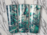 Three tumblers with iridescent teal and white butterfly and swirl designs on marble.